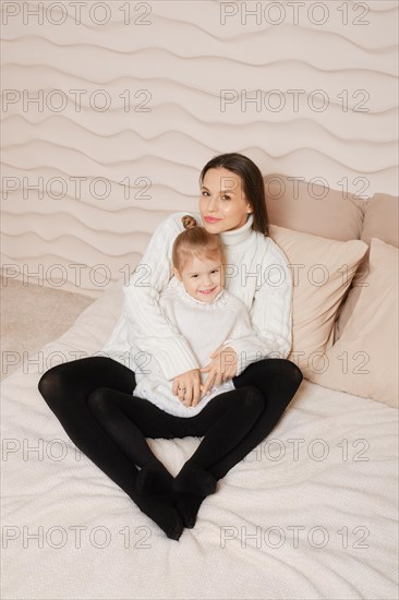 Family photo of pretty mother with long dark hair wearing knitted white sweater posing with her little cute daughter in cozy bed