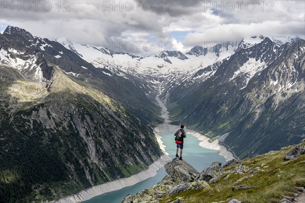 Mountaineer on a rock in front of a mountain panorama, view of Schlegeisspeicher, glaciated rocky mountain peaks Hoher Weisszint and Hochfeiler with glacier Schlegeiskees, Berliner Hoehenweg, Zillertal Alps, Tyrol, Austria, Europe