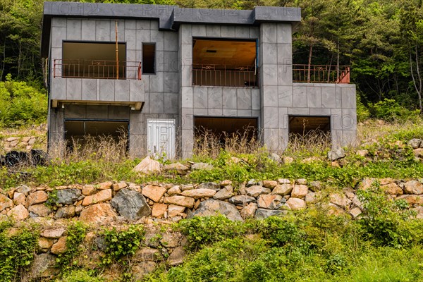 Unfinished abandoned gray concrete house in the side of a hill with stone wall in the foreground and lush green foliage in the background