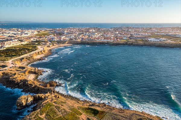 Drone view of waves break on rocky shores in town of Peniche, Portugal. Summer sunset haze, little foliage and rocky cliffs, peninsula and rocks, fishing town, horizon