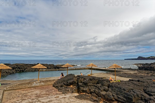 Natural swimming pool by the sea with parasols and people relaxing, lava rocks coastal walk Ponta da Iiha, Calhau, west coast, Pico, Azores