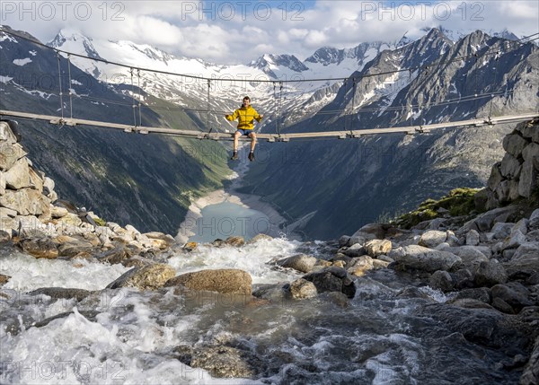 Mountaineer sitting on a suspension bridge over a mountain stream Alelebach, picturesque mountain landscape near the Olpererhuette, view of turquoise blue lake Schlegeisspeicher, glaciated rocky mountain peaks Hoher Weisszint and Hochfeiler with glacier Schlegeiskees, Berliner Hoehenweg, Zillertal Alps, Tyrol, Austria, Europe