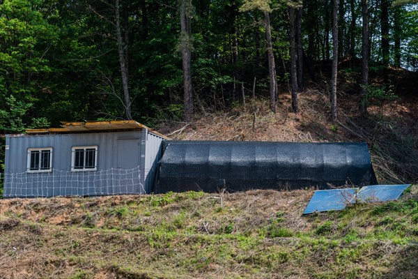 Two storage buildings on hillside in wooded farm region, one metal and one support frame covered with black mesh
