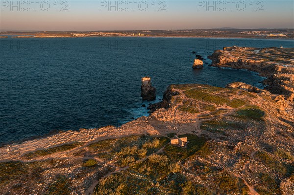 Bird's-eye view of sunset over ocean waves break on rocky shores, Peniche, Portugal. Summer sunset haze, little foliage and rocky cliffs, peninsula and rocks, fishing town, horizon