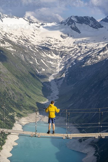 Mountaineers on a suspension bridge over a mountain stream Alelebach, picturesque mountain landscape near the Olpererhuette, view of turquoise-blue lake Schlegeisspeicher, glaciated rocky mountain peaks Grosser Moeseler, Hoher Weisszint and Hochfeilermit glacier Schlegeiskees, Berliner Hoehenweg, Zillertal Alps, Tyrol, Austria, Europe