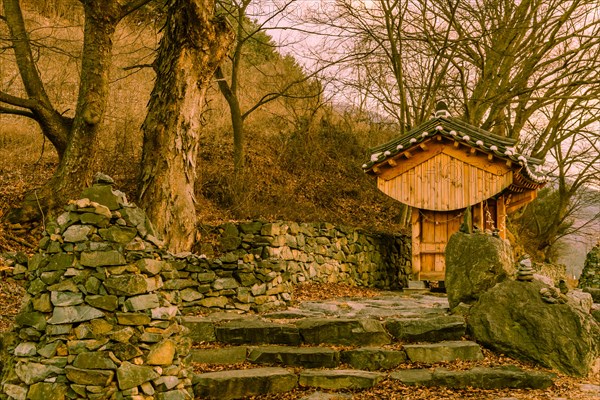 Small wooden oriental building with tiled roof in front of rock wall at rural roadside park on winter evening