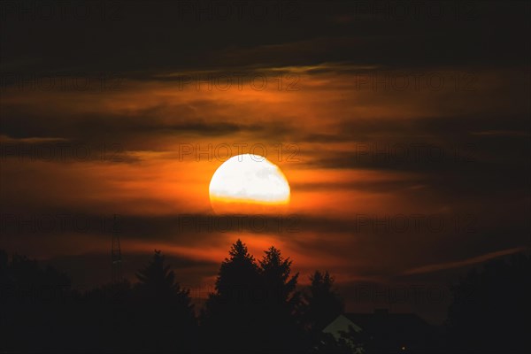 An atmospheric moonrise with orange glowing clouds and tree silhouettes, Haan, North Rhine-Westphalia, Germany, Europe