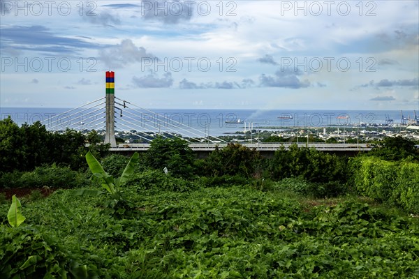 A highway with Mauritian emblem on pillar and a truck driving, surrounded by mountains and an overcast sky.A1-M1 Link Bridge At Grand River North West Valley on the island of Mauritius