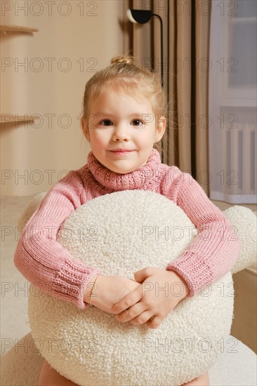 In a cozy room, little girl in pink sweater embraces a large white stuffed decorative pillow