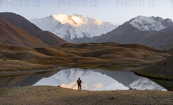 Hiker looking at mountain landscape, mountains reflected in a small mountain lake, Lenin Peak, Trans Alay Mountains, Pamir Mountains, Osh Province, Kyrgyzstan, Asia