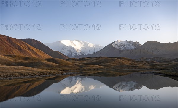 White glaciated and snowy mountain peak Pik Lenin at sunset, mountains reflected in a lake between golden hills, Trans Alay Mountains, Pamir Mountains, Osh Province, Kyrgyzstan, Asia