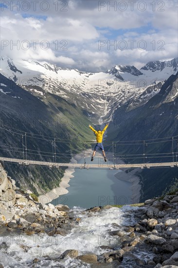 Mountaineer jumping on a suspension bridge over a mountain stream Alelebach, picturesque mountain landscape near the Olpererhuette, view of turquoise-blue lake Schlegeisspeicher, glaciated rocky mountain peaks Grosser Moeseler, Hoher Weisszint and Hochfeiler with glacier Schlegeiskees, Berliner Hoehenweg, Zillertal Alps, Tyrol, Austria, Europe