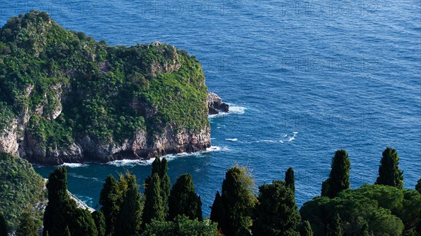 Cliff on a coast with lush greenery surrounded by the blue sea with visible waves, Taormina, Eastern Sicily, Sicily, Italy, Europe