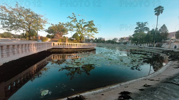A serene urban pond called Srah Chhouk or Lotus Pond in the middle of Kampot town with reflected trees under a blue sky at sunset. Kampot, Cambodia, Asia