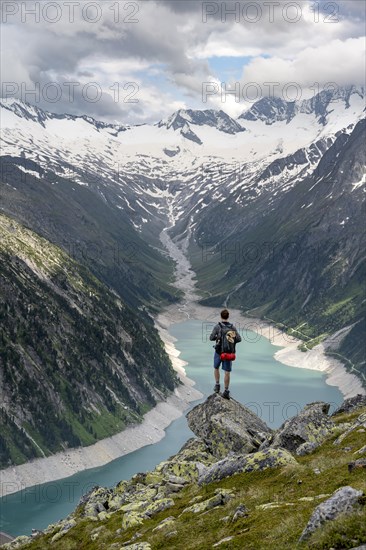 Mountaineer on a rock in front of a mountain panorama, view of Schlegeisspeicher, glaciated rocky mountain peaks Hoher Weisszint and Hochfeiler with glacier Schlegeiskees, Berliner Hoehenweg, Zillertal Alps, Tyrol, Austria, Europe