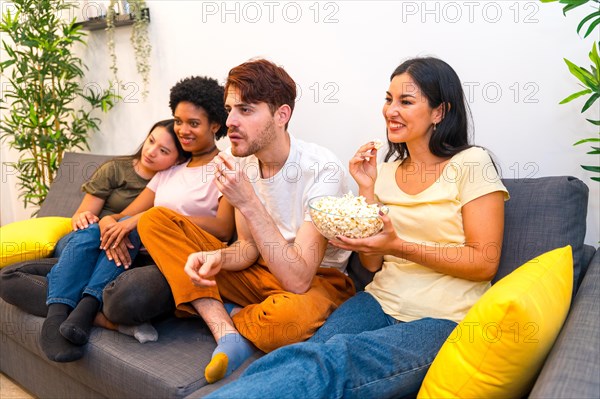 Flatmates watching an interesting movie sitting together on the sofa paying attention and eating pop corn