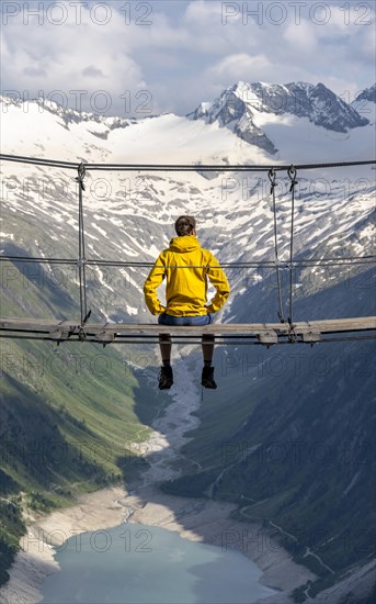 Mountaineer sitting on a suspension bridge, picturesque mountain landscape near the Olpererhuette, view of turquoise-blue lake Schlegeisspeicher, glaciated rocky mountain peaks Hoher Weisszint and Hochfeiler with glacier Schlegeiskees, Berliner Hoehenweg, Zillertal Alps, Tyrol, Austria, Europe