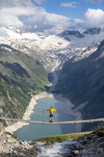 Mountaineers on a suspension bridge over a mountain stream Alelebach, picturesque mountain landscape near the Olpererhuette, view of turquoise-blue lake Schlegeisspeicher, glaciated rocky mountain peaks Grosser Moeseler, Hoher Weisszint and Hochfeiler with glacier Schlegeiskees, Berliner Hoehenweg, Zillertal Alps, Tyrol, Austria, Europe