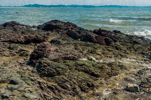 Seascape of rocking shore with beach and harbor road in background under cloudy blue sky