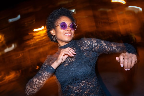 Night photo with flash and motion of an african woman with sunglasses dancing in the city street at night