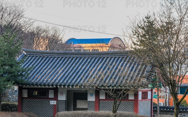 Public restroom in woodland park with a tiled roof surrounded by trees and brushes with a modern building with a blue roof in the background