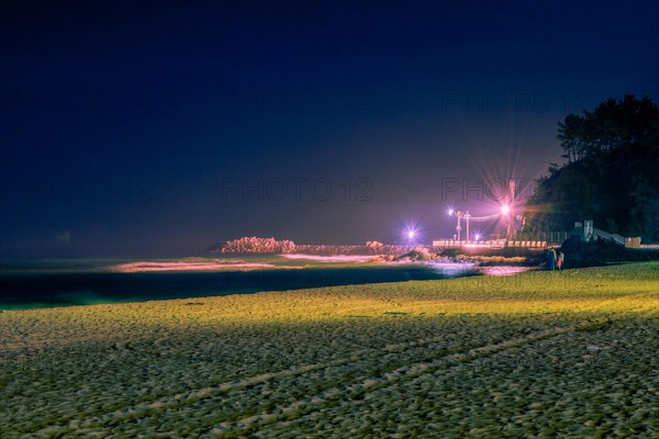 Night scene at beach bathed in soft light with unrecognizable people and pier lit up by street lights in background
