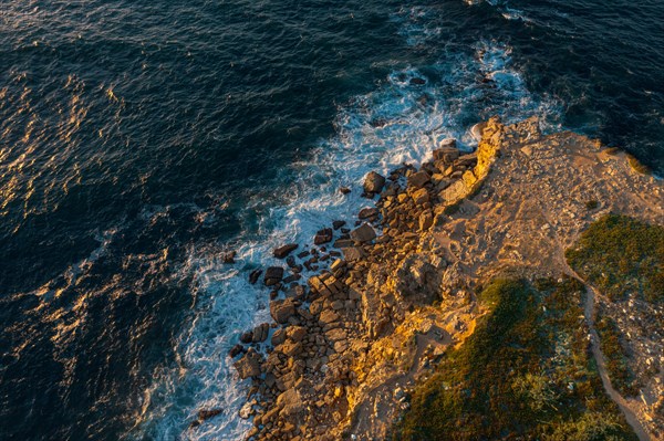 Drone top down view of ocean waves break on rocky headland. Drone shot of sea blue waves beat and splash in summer sunset haze, little foliage and rocky cliffs, establishing or static shot, Peniche, Portugal, Europe