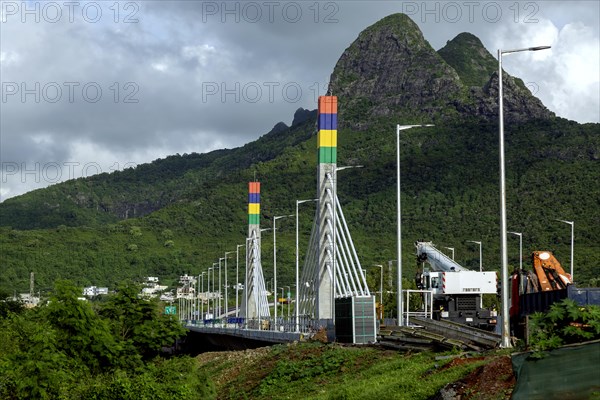 A highway with Mauritian emblem on pillar and a truck driving, surrounded by mountains and an overcast sky.A1-M1 Link Bridge At Grand River North West Valley on the island of Mauritius