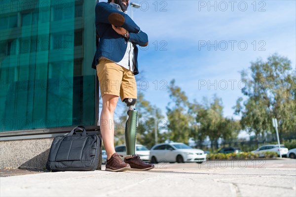 Businessman with prosthetic leg waiting patiently leaning on a building with arms crossed in the street