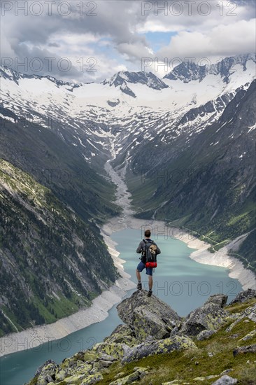 Mountaineer on a rock in front of a mountain panorama, view of Schlegeisspeicher, glaciated rocky mountain peaks Hoher Weisszint and Hochfeiler with glacier Schlegeiskees, Berliner Hoehenweg, Zillertal Alps, Tyrol, Austria, Europe