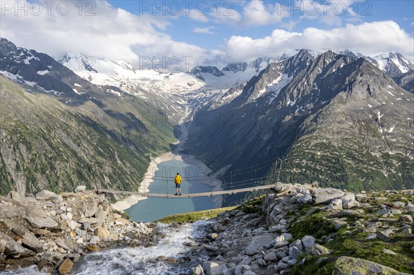 Mountaineers on a suspension bridge over a mountain stream Alelebach, picturesque mountain landscape near the Olpererhuette, view of turquoise-blue lake Schlegeisspeicher, glaciated rocky mountain peaks Grosser Moeseler, Hoher Weisszint and Hochfeiler with glacier Schlegeiskees, Berliner Hoehenweg, Zillertal Alps, Tyrol, Austria, Europe