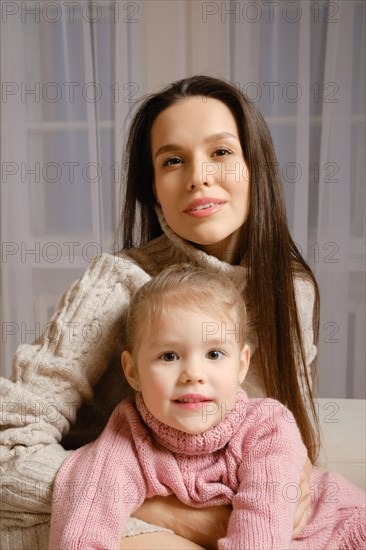 A beautiful and loving mother and a sweet daughter are sitting together in the living room in knitted sweaters