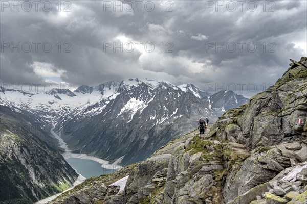 Two mountaineers on hiking trail with snow, view of Schlegeisspeicher, glaciated rocky mountain peaks Hoher Weisszint and Hochfeiler with glacier Schlegeiskees, Berliner Hoehenweg, Zillertal Alps, Tyrol, Austria, Europe