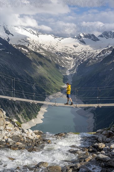 Mountaineers on a suspension bridge over a mountain stream Alelebach, picturesque mountain landscape near the Olpererhuette, view of turquoise-blue lake Schlegeisspeicher, glaciated rocky mountain peaks Grosser Moeseler, Hoher Weisszint and Hochfeiler with glacier Schlegeiskees, Berliner Hoehenweg, Zillertal Alps, Tyrol, Austria, Europe