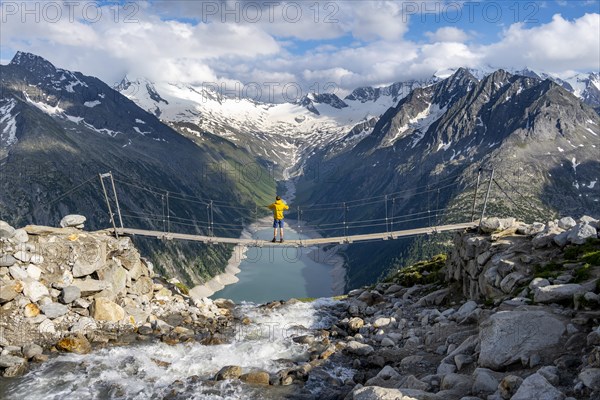 Mountaineers on a suspension bridge over a mountain stream Alelebach, picturesque mountain landscape near the Olpererhuette, view of turquoise-blue lake Schlegeisspeicher, glaciated rocky mountain peaks Hoher Weisszint and Hochfeiler with glacier Schlegeiskees, Berliner Hoehenweg, Zillertal Alps, Tyrol, Austria, Europe