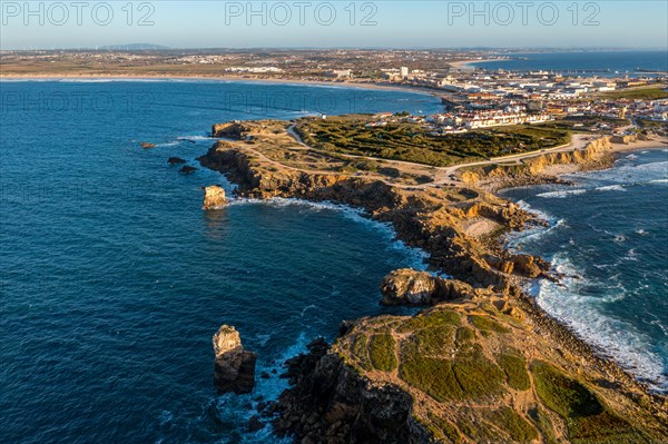 Aerial image of peninsula and town of Peniche, Portugal on sunny day. Summer sunset haze, little foliage and rocky cliffs, peninsula and rocks, fishing town, horizon, ocean waves break on rocky shores