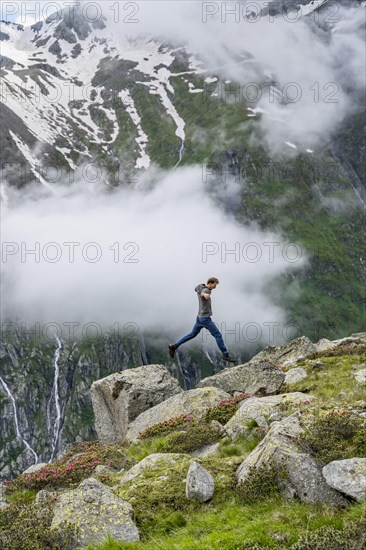 Mountaineer jumping from one rock to the next, cloudy mountain landscape with blooming alpine roses, view of rocky and glaciated mountains, Furtschaglhaus, Berliner Hoehenweg, Zillertal, Tyrol, Austria, Europe