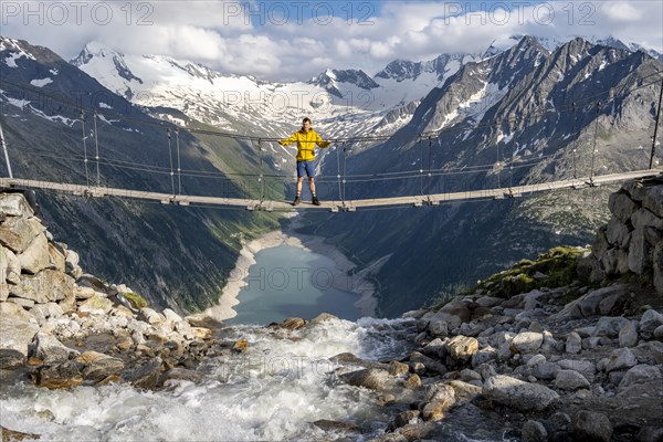 Mountaineers on a suspension bridge over a mountain stream Alelebach, picturesque mountain landscape near the Olpererhuette, view of turquoise-blue lake Schlegeisspeicher, glaciated rocky mountain peaks Grosser Moeseler, Hoher Weisszint and Hochfeiler with glacier Schlegeiskees, Berliner Hoehenweg, Zillertal Alps, Tyrol, Austria, Europe