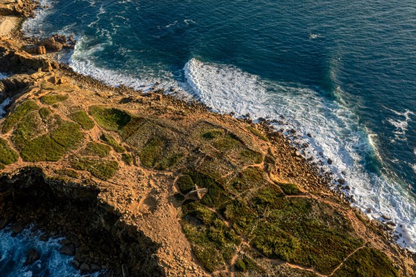 Ocean waves break on rocky island shores at sunset. Drone shot of sea blue waves beat and splash in summer sunset haze, little foliage and rocky cliffs, establishing or static shot, Peniche, Portugal, Europe