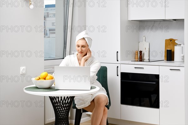 Serious young woman in bathrobe sits in the kitchen with laptop