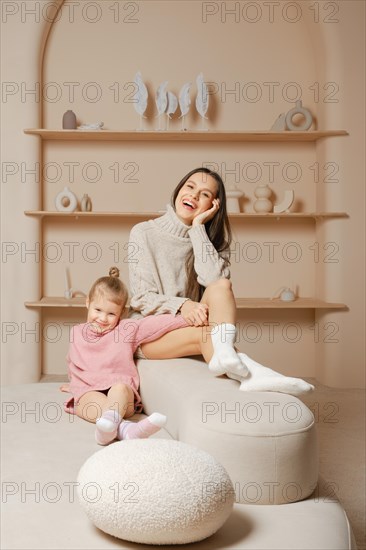 Young mother and little daughter share a funny moment on a couch in a well-lit living room. The daughter wears a pink sweater, and the mother wears a white sweater