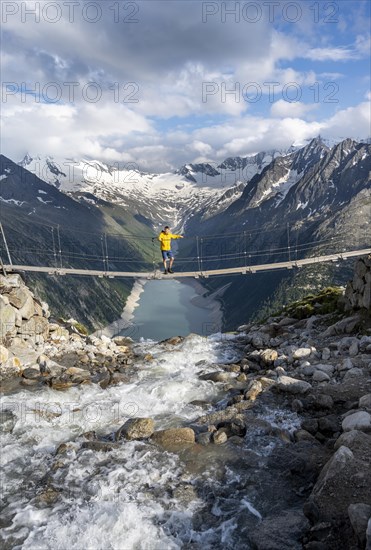 Mountaineers on a suspension bridge over a mountain stream Alelebach, picturesque mountain landscape near the Olpererhuette, view of turquoise-blue lake Schlegeisspeicher, glaciated rocky mountain peaks Hoher Weisszint and Hochfeiler with glacier Schlegeiskees, Berliner Hoehenweg, Zillertal Alps, Tyrol, Austria, Europe