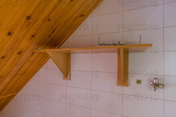 Water faucet on white tiled wall next to wooden shelf in laundry room of abandoned house