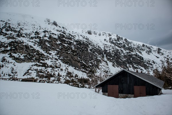 A solitary cabin sits beneath a rugged, snowy mountain landscape under an overcast sky. Poland, Karpacz, Karkonosze