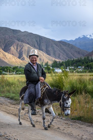 Man in traditional dress riding a donkey in front of a mountain backdrop, Kyrgyzstan, Asia