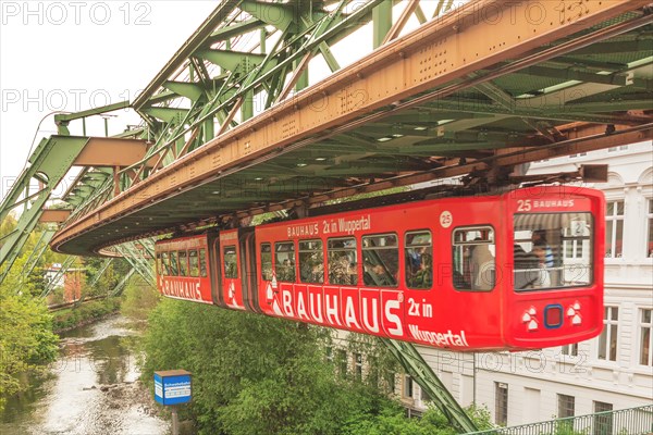 Red suspension railway with advertising runs over an urban river landscape, suspension railway, Wuppertal, North Rhine-Westphalia, Germany, Europe