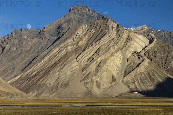 Impressive cliff in the Zanskar Mountains of the Himalayas, with clearly visible folded strata, the rock layers of sediment or metamorphic rocks, and Rangdum Gompa, the Buddhist monastery located below the rock face, on a small hill above the valley floor. Photographed in September, the late summer, at moonrise of a clear afternoon, with the moon in view in the blue sky. Kargil District, Union Territory of Ladakh, India, Asia