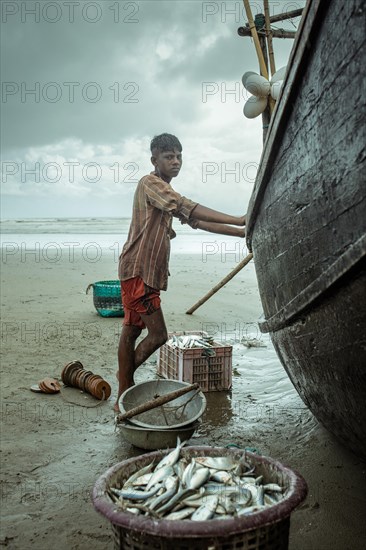 Fisherman by his boat on the beach with the catch of the day during a monsoon shower, Cox's Bazar, Bangladesh, Asia