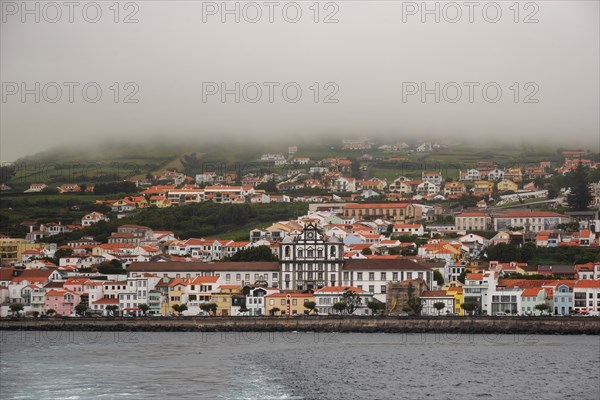 The coastal town of Horta under the fog, colourful houses nestle against green hills with calm sea in the foreground, Horta, Faial, Azores, Portugal, Europe