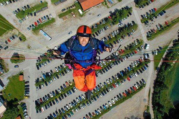 Paragliding pilot with orange and blue clothes, flying high over parking lot of Brauneck cable car, bird's eye view, Lenggries, Bavaria, Germany, Europe